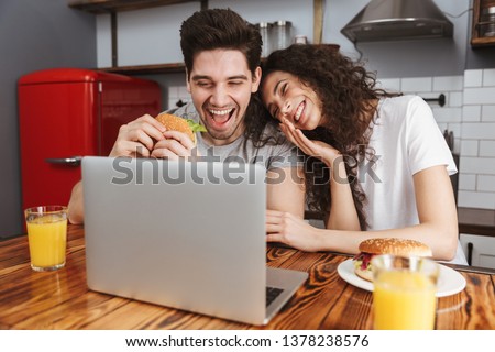 Picture of positive couple man and woman 30s looking at laptop on table while eating hamburger in kitchen at home