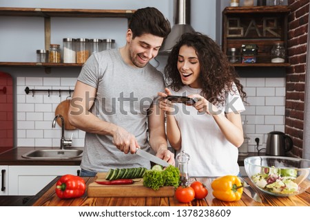 Picture of adorable couple man and woman 30s cooking salat with vegetables together in modern kitchen at home