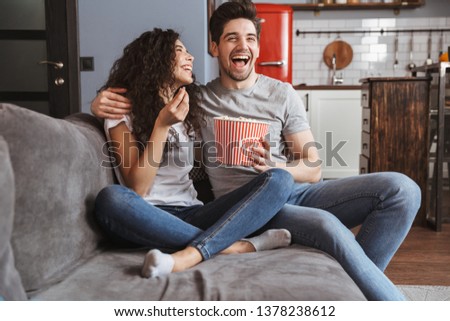 Picture of joyful young couple man and woman 30s sitting on sofa at home and eating popcorn from bucket