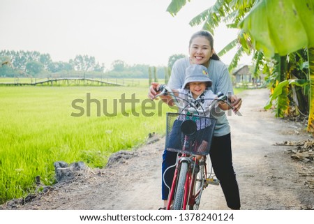 Young mother teaching her son how to ride bicycle at nature with copy space for your text. Concept of friendly family and of summer vacation. Paddy farm plant peaceful.