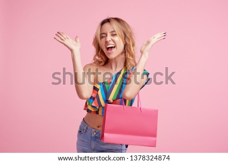 Portrait of an excited beautiful girl wearing colorful clothes holding shopping bag isolated over pink background.  Place to sign or text  on the package