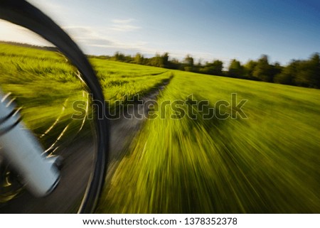 Blurred background in motion: a bicycle wheel rides along a dirt road in the fields with fresh green beautiful grass against a blue sky on a bright sunny day. Activities and sports
