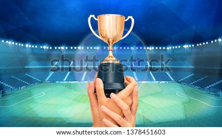 Hands hold up trophy with background football stadium.