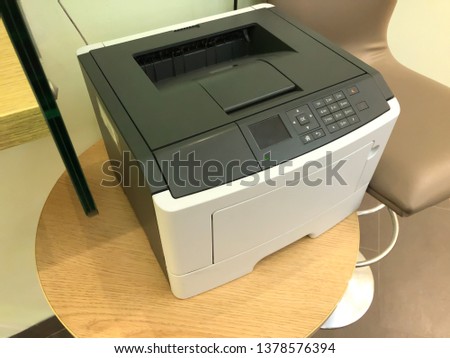 Office laser printer on a small table next to the office chair connected to the network for shared printing from many devices and computers. Printer with numeric keypad and function screen.