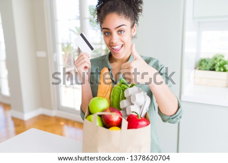 Young african american girl holding paper bag of groceries and credit card as payment happy with big smile doing ok sign, thumb up with fingers, excellent sign