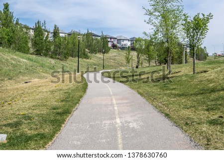 Paved bike path through a grass field lined with trees on a cloudy summer day. 
