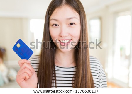 Beautiful Asian woman holding credit card with a happy face standing and smiling with a confident smile showing teeth