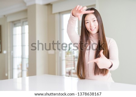 Beautiful Asian woman wearing casual sweater on white table smiling making frame with hands and fingers with happy face. Creativity and photography concept.