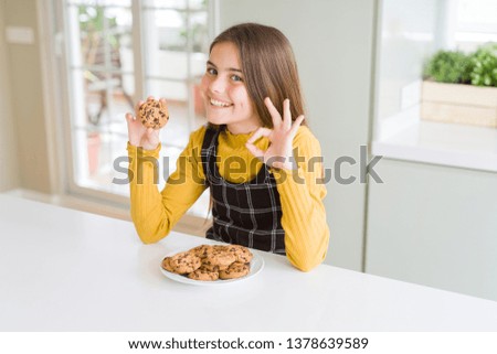 Beautiful young girl kid eating chocolate chips cookies doing ok sign with fingers, excellent symbol