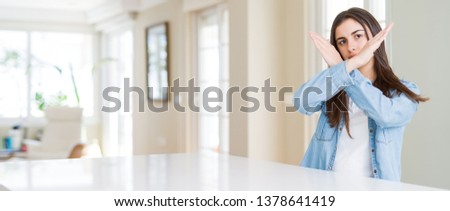 Wide angle picture of beautiful young woman sitting on white table at home Rejection expression crossing arms doing negative sign, angry face