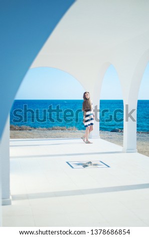Image of caucassian girl in striped dress with long brown hair looking at the beach in the distance, at the blue sea. Warm summer day. White arch and columns of Ayia Thekla Chapel