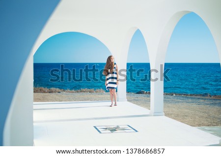 Image of caucassian girl in striped dress with long brown hair looking at the beach in the distance, at the blue sea. Warm summer day. White arch and columns of Ayia Thekla Chapel