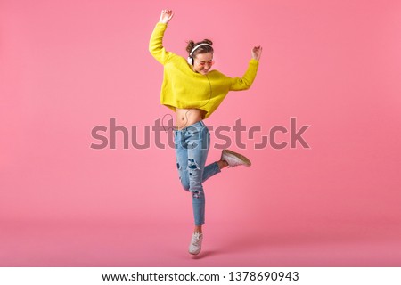 attractive happy funny woman jumping listening to music in headphones dressed in hipster colorful style outfit isolated on pink studio background, wearing yellow sweater and sunglasses, having fun