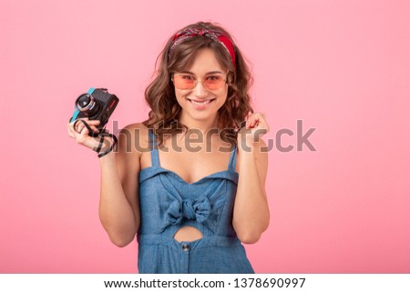 attractive smiling woman taking photo on vintage camera wearing denim dress and sunglasses isolated on pink background, traveler on vacation, summer fashion style