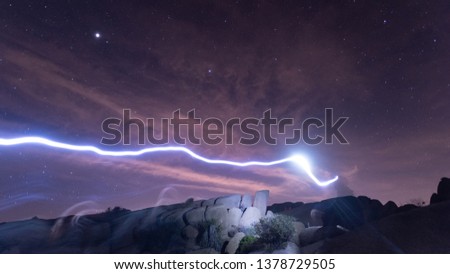 long exposure image of man with headlight leaving a light streak with desert rock formations in the background under a starry night sky in Joshua Tree National Park in California