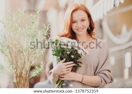 Outdoor shot of pleasant looking young ginger female with satisfied expression, wears brown jumper, holds bouquet of white flowers, walks on street against blurred background. Sunny spring day