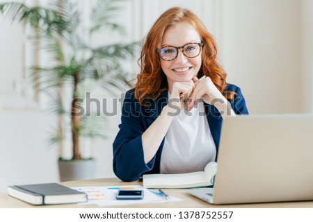 Pleased cheerful red haird female economist develops financial startup project, poses in office interior, works in business sphere, dressed in formal clothes, has happy expression, owns company Royalty-Free Stock Photo #1378752557