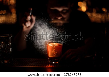 Darkened picture of male bartender with beard in gray shirt pours an alcohol cocktail using steel sprayer