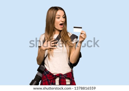 Young photographer woman holding a credit card and surprised on isolated blue background