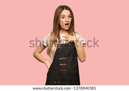 Young woman with apron surprised and shocked while looking right on isolated pink background