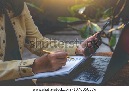 Casual young woman hand using mobile smart phone, writing on blank notebook with laptop computer and cup of coffee on the table at outdoor nature background. Online studying, e learning concept.