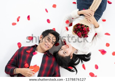 Top view of happy young couple looking at each other and smiling while lying on white background floor. They is holding a red paper heart, and Rose petals fall full on the White Background