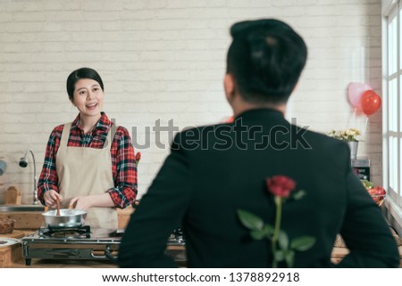 Little surprise for her. Rear view of man holding a red rose behind his back while his girlfriend standing on background smiling cooking on stove on valentine day. husband send wife flower.