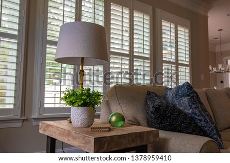 Beautiful light wooden side table with bright green leaves, oil lamp, and lamp on it. A neutral colored sofa is next to it, with shuttered windows behind it. Beautiful interior design. Royalty-Free Stock Photo #1378959410
