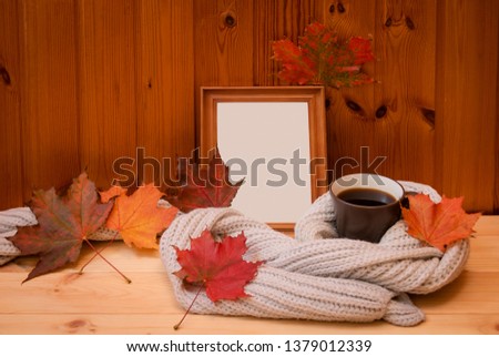 Empty wooden frame, mug of coffee wrapped in gray woolen scarf and colorful autumn leaves on wooden table. View with copy space.