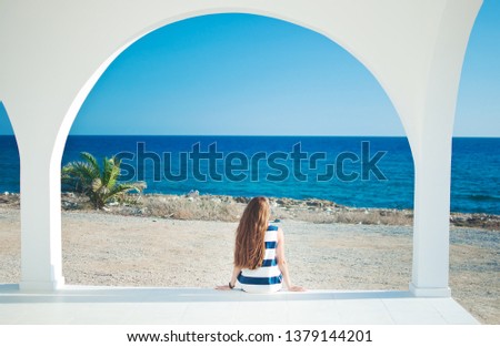 Image of a back of one caucassian girl in striped dress with long brown hair looking at the beach in the distance, at the blue sea. Warm summer day. White arch and columns of Ayia Thekla Chapel