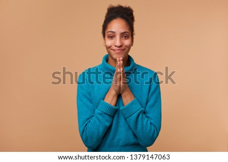 Portrait of young African American girl with curly dark hair wearing a blue sweater. Smiling, keeps palms together, pleads for mercy. Isolated over beige background.