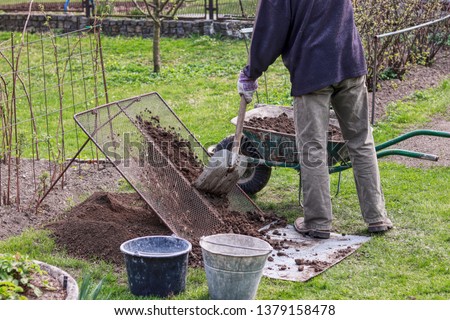 Man wears work outfit. Gardener prepare earth for new flowers and trees. He sifting soil through handmade sieve for better consistency earth and without stones. Plastic black bucket for stones Royalty-Free Stock Photo #1379158478