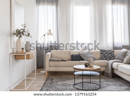 Round coffee table in front of beige sofa with pillows in bright living room interior with console table with flowers in vase