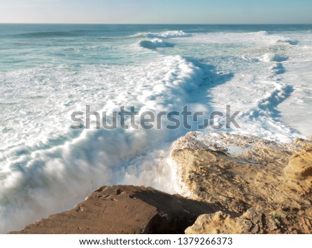 Empty long wide beach, yellow sand; curling crushing waves ; sunny weather in Nazare, Portugal; nature background; windy and wavy beach with surf camp; nature power; dangerous cliff