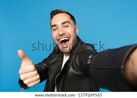 Close up selfie of stylish young unshaven man in black leather jacket white t-shirt looking camera isolated on blue background studio portrait. People sincere emotions concept. Mock up copy space