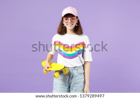 Portrait of smiling teen girl in vivid clothes, heart eyeglasses holding yellow skateboard isolated on violet pastel wall background. People sincere emotions, lifestyle concept. Mock up copy space