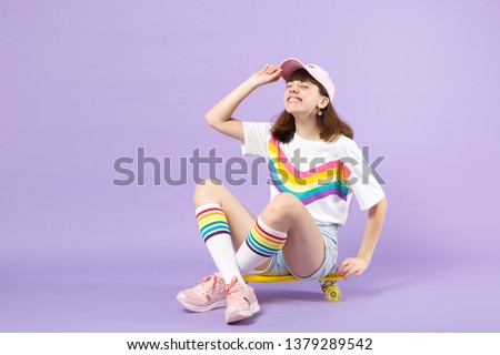 Happy teen girl in vivid clothes sitting on yellow skateboard, keeping eyes closed isolated on violet pastel wall background in studio. People sincere emotions lifestyle concept. Mock up copy space