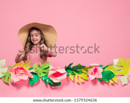 Portrait of a little girl in a summer hat on a pink background with paper flowers, place for text, summer advertising concept