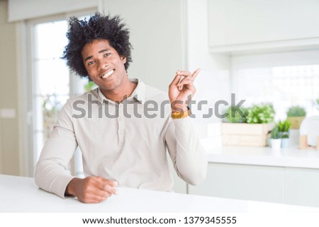 African American man at home with a big smile on face, pointing with hand and finger to the side looking at the camera.
