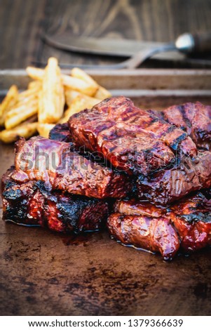 Bbq boneless beef ribs with barbecue sauce and potato wedges over a rustic background. Extreme shallow depth of field with blurred background and selective focus on front of meat.