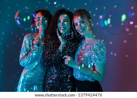 Happy Three beauty women wearing in shiny clothes posing together with soap bubbles and looking at the camera over sparkling background