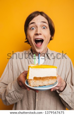 Vertical image of Surprised happy handsome man in shirt holding plate with cake and looking at the camera over yellow background