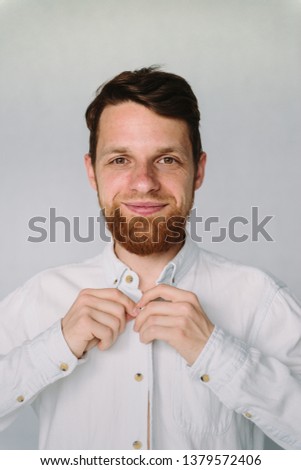 Young hipster man with beard buttoning white shirt, looking to camera and smiling. Vertical shot of smiling happy hipster businessman with beard  in white casual shirt. Happy man on white background