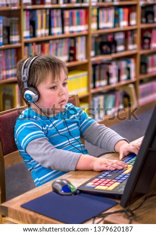 A little boy wearing headphones, plays on a lap top at the local public library