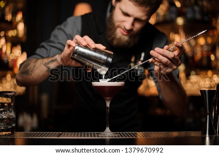 Young tattooed male bartender with beard makes red alcohol cocktail using steel shaker and long bar spoon at bar counter Royalty-Free Stock Photo #1379670992