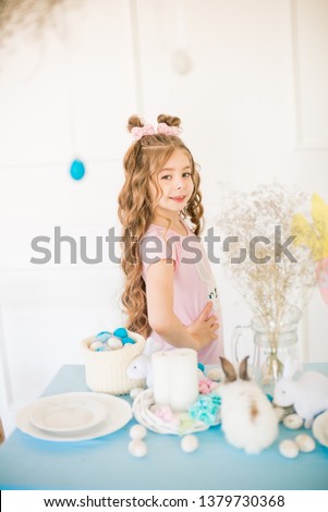 Little cute girl with long curly hair with little bunnies and Easter decor at home at the holiday table.