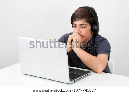 Caucasian teenage boy wearing wireless headphones sitting at the desk looking at the laptop screen. Concept of modern education.