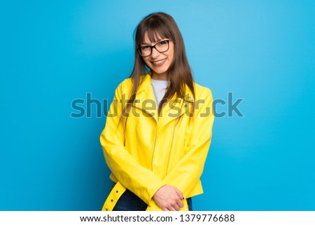 Young woman with yellow jacket on blue background with glasses and happy