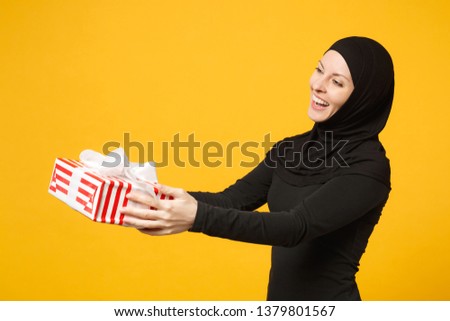Smiling young arabian muslim woman in hijab black clothes hold in hand present box with gift isolated on yellow wall background studio portrait. People religious lifestyle concept. Mock up copy space