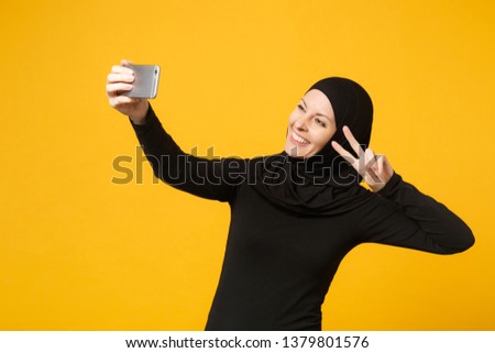 Smiling young arabian muslim woman in hijab black clothes doing selfie shot on mobile phone isolated on yellow wall background, studio portrait. People religious lifestyle concept. Mock up copy space